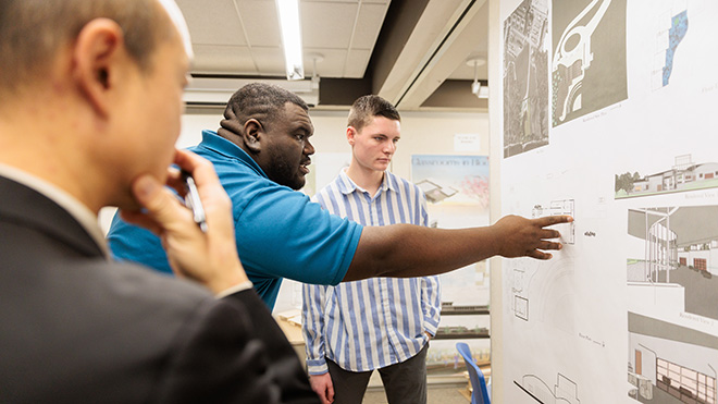 An Architecture alum points to a spot on a poster as a student stands next to him