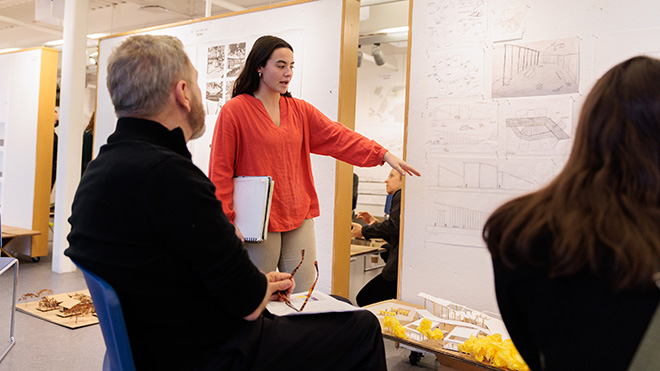 An Architecture student points to a spot on a poster showing her design as two reviewers sit and listen